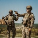 Two U.S. Army soldiers in full combat gear are standing on elevated gravel terrain, with one soldier pointing in a direction while the other carries equipment. Both are wearing Operational Camouflage Pattern (OCP) uniforms, plate carriers, and helmets. The background shows a wide open training range with vegetation and what appear to be military vehicles in the distance.