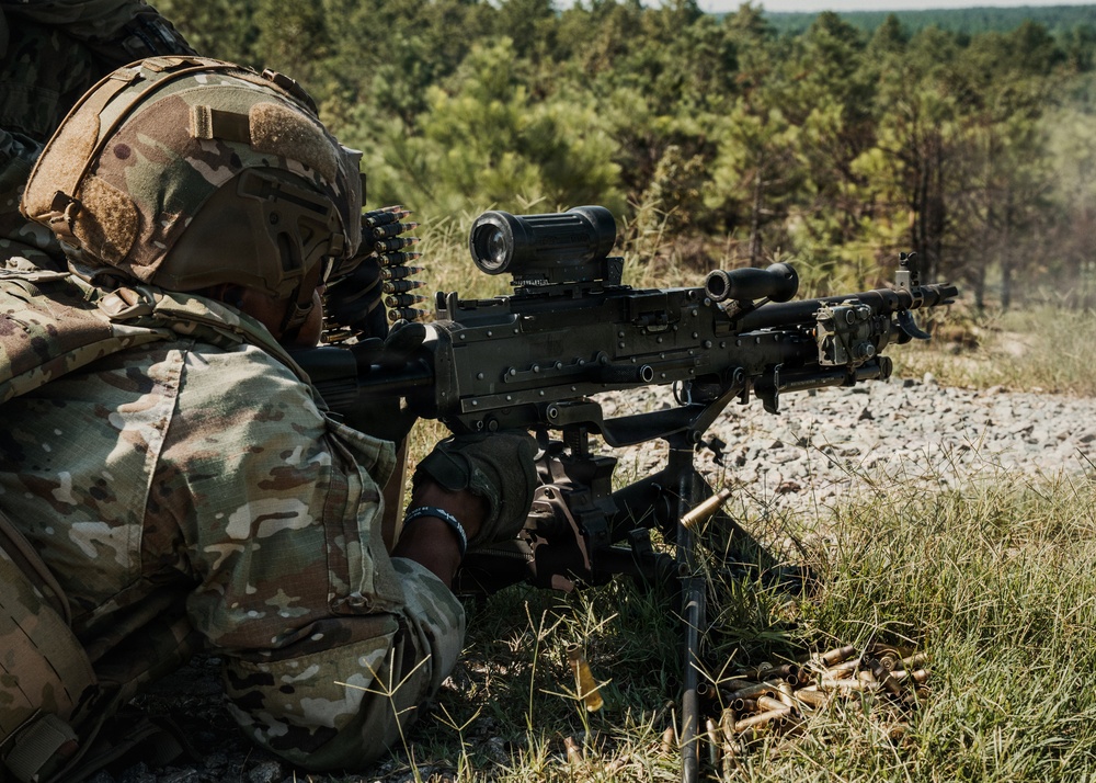 A soldier in full multicam camouflage gear is in a prone/low firing position operating a belt-fed machine gun mounted on a bipod. Spent brass casings are visible on the ground, indicating the weapon has been fired. The setting appears to be a wooded training range or field environment.