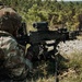 A soldier in full multicam camouflage gear is in a prone/low firing position operating a belt-fed machine gun mounted on a bipod. Spent brass casings are visible on the ground, indicating the weapon has been fired. The setting appears to be a wooded training range or field environment.