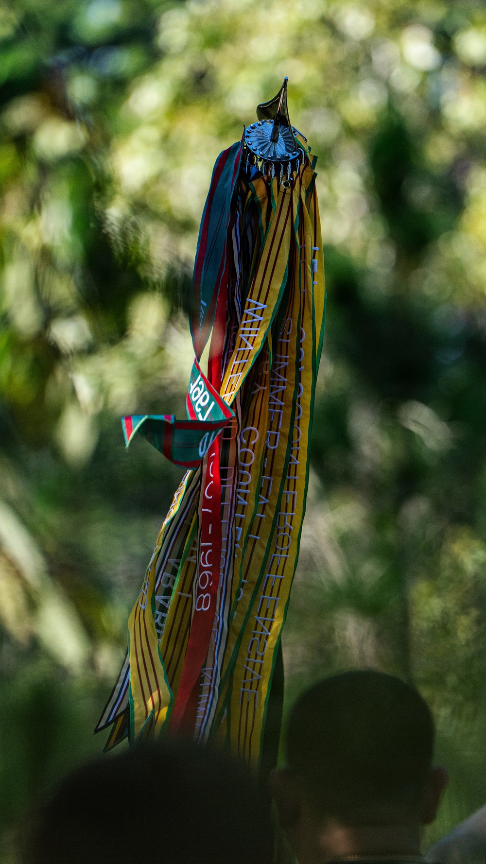 The image shows a ceremonial or decorative staff/pole topped with a dark metal ornament, from which hang multiple colorful ribbons or sashes. The ribbons appear to bear printed text including partially readable words such as 'SUMMER CONFERENCE' and a date that appears to include '1968'. The background is a heavily blurred green foliage setting, suggesting an outdoor environment.