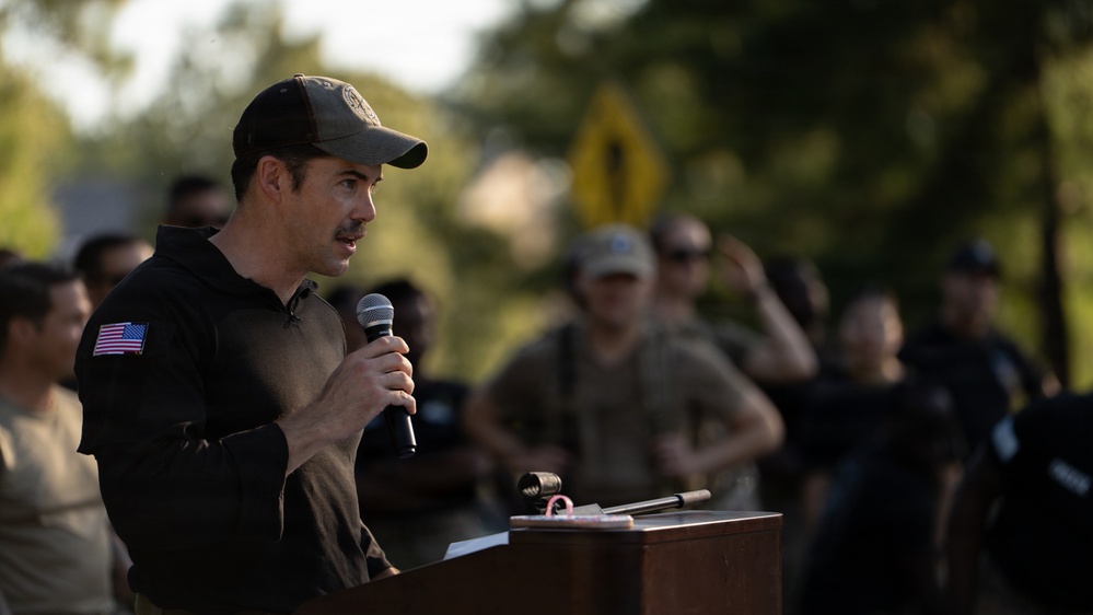 A man wearing a dark tactical-style jacket with a US flag patch on the sleeve and a baseball cap is speaking into a microphone at a wooden podium. He appears to be addressing a group of individuals in the background who are dressed in military or tactical attire. The setting appears to be an outdoor event, likely a military or law enforcement gathering.
