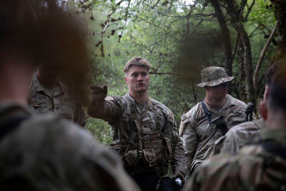 A group of U.S. Army soldiers are gathered in a wooded outdoor environment, with one soldier appearing to brief or instruct the others using hand gestures. The soldiers are wearing OCP (Operational Camouflage Pattern) uniforms and tactical gear. The setting suggests a field training exercise or operational briefing in a forested area.