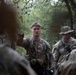 A group of U.S. Army soldiers are gathered in a wooded outdoor environment, with one soldier appearing to brief or instruct the others using hand gestures. The soldiers are wearing OCP (Operational Camouflage Pattern) uniforms and tactical gear. The setting suggests a field training exercise or operational briefing in a forested area.
