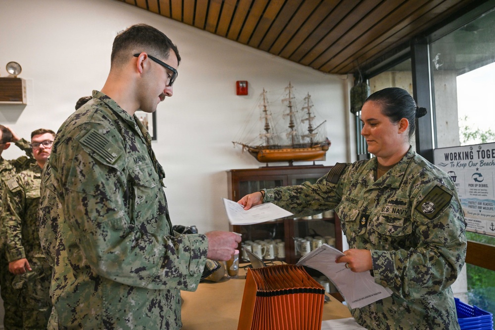 Two U.S. Navy personnel in NWU Type III camouflage uniforms are exchanging documents at an indoor table. A female sailor with 'U.S. NAVY' tape visible is handing paperwork to a male sailor wearing glasses. The setting appears to be a visitor center or administrative building, with a model tall ship and environmental signage visible in the background.