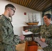 Two U.S. Navy personnel in NWU Type III camouflage uniforms are exchanging documents at an indoor table. A female sailor with 'U.S. NAVY' tape visible is handing paperwork to a male sailor wearing glasses. The setting appears to be a visitor center or administrative building, with a model tall ship and environmental signage visible in the background.