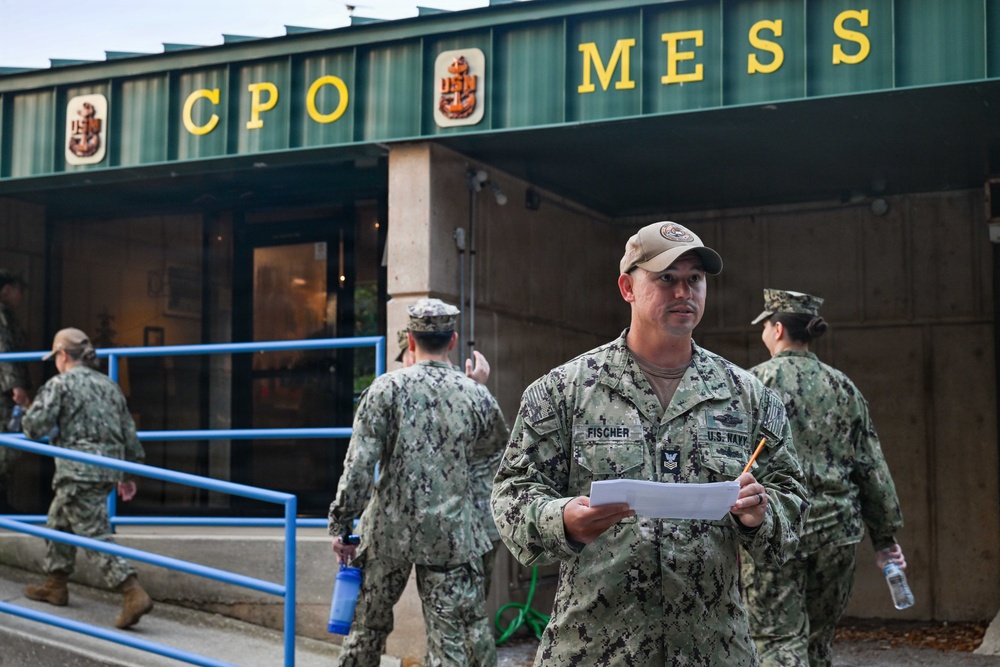 The image shows U.S. Navy personnel in NWU Type III (woodland camouflage) uniforms gathered outside a CPO Mess (Chief Petty Officers' dining facility). A sailor identified by name tape as 'FISCHER' stands in the foreground holding papers and a pencil, appearing to be coordinating or checking personnel. The setting appears to be a naval installation, likely during a muster or organized event.