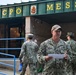 The image shows U.S. Navy personnel in NWU Type III (woodland camouflage) uniforms gathered outside a CPO Mess (Chief Petty Officers' dining facility). A sailor identified by name tape as 'FISCHER' stands in the foreground holding papers and a pencil, appearing to be coordinating or checking personnel. The setting appears to be a naval installation, likely during a muster or organized event.