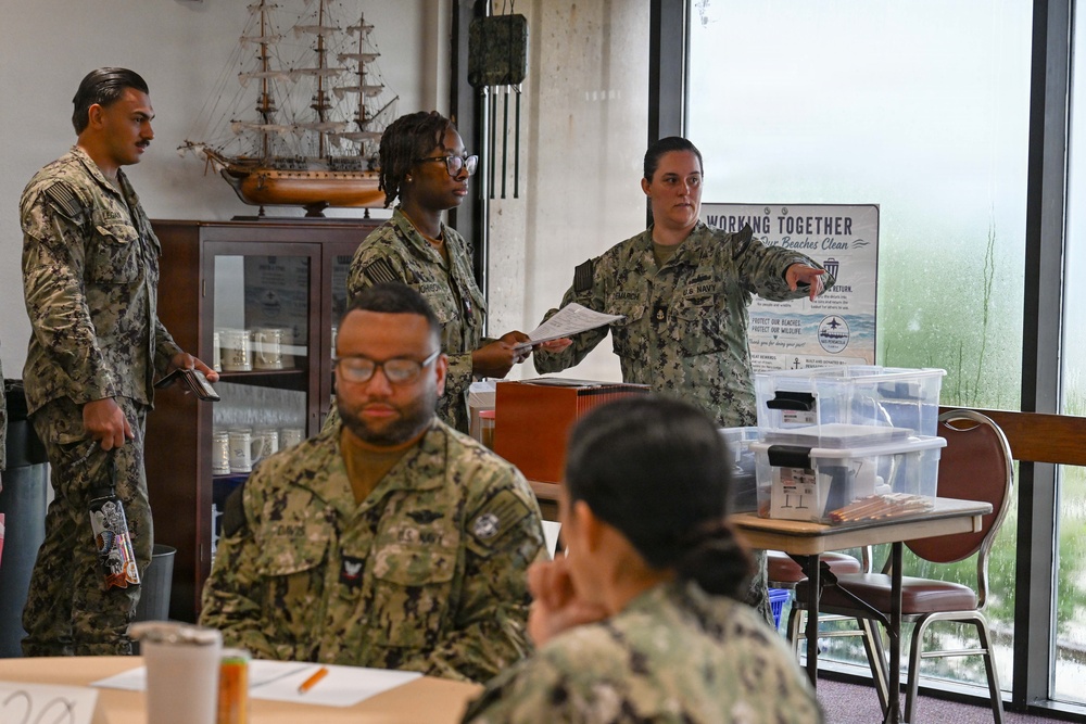 A group of U.S. Navy personnel in NWU (Navy Working Uniform) camouflage are gathered in what appears to be a briefing or administrative room. One female sailor appears to be directing or instructing others, holding a document and pointing. The setting includes a ship model display cabinet, a 'Working Together' environmental poster, and a table with clear plastic storage containers.