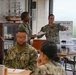 A group of U.S. Navy personnel in NWU (Navy Working Uniform) camouflage are gathered in what appears to be a briefing or administrative room. One female sailor appears to be directing or instructing others, holding a document and pointing. The setting includes a ship model display cabinet, a 'Working Together' environmental poster, and a table with clear plastic storage containers.