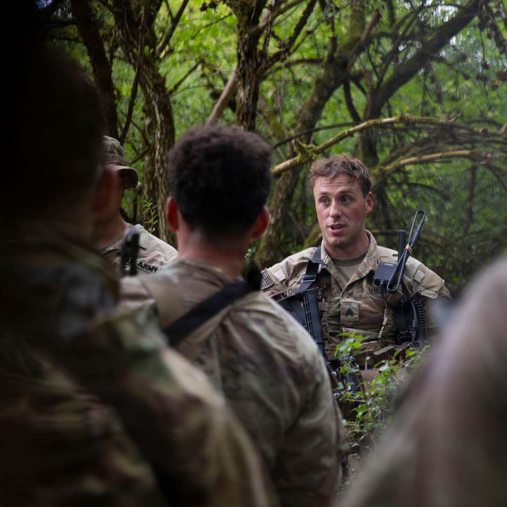 A U.S. Army Sergeant in MultiCam/OCP camouflage uniform is addressing a small group of soldiers in a dense forested environment. The sergeant appears to be conducting a briefing or after-action review during a field training exercise. The setting suggests a woodland training area with heavy tree cover.
