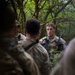 A U.S. Army Sergeant in MultiCam/OCP camouflage uniform is addressing a small group of soldiers in a dense forested environment. The sergeant appears to be conducting a briefing or after-action review during a field training exercise. The setting suggests a woodland training area with heavy tree cover.