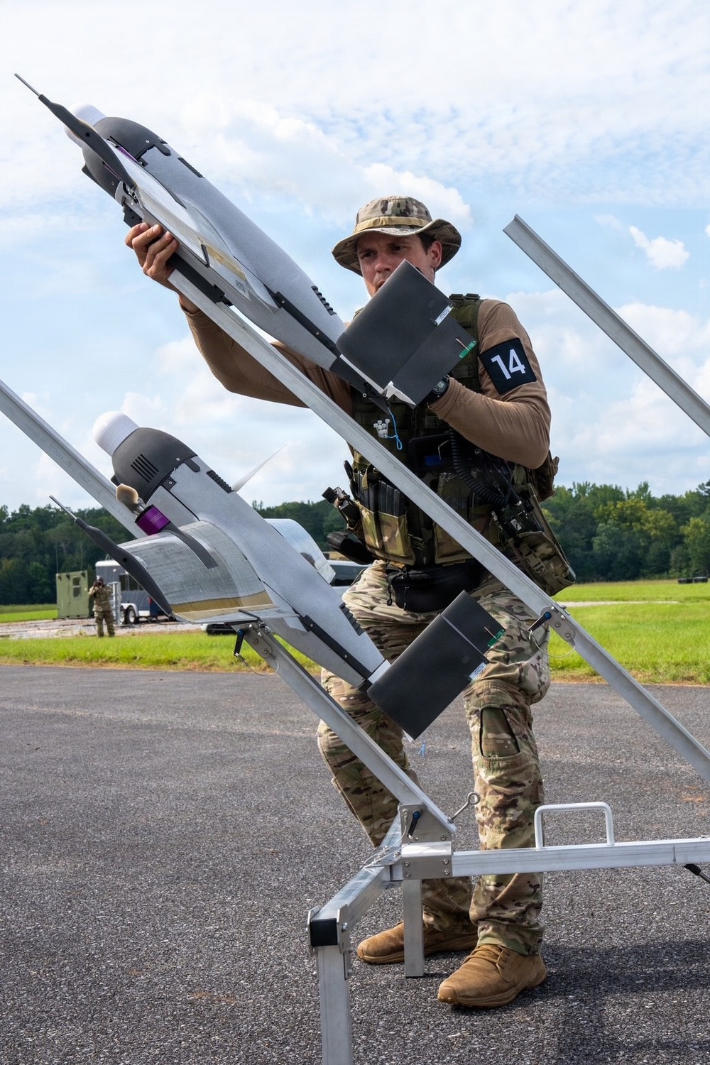 A military personnel member wearing multicam camouflage uniform and a boonie hat is loading or positioning what appears to be a fixed-wing UAV (unmanned aerial vehicle) onto an angled launch rail system. The operator wears a tactical vest and has an armband marked with the number '14'. The setting appears to be a paved airfield or tarmac area with green grass and trees visible in the background.