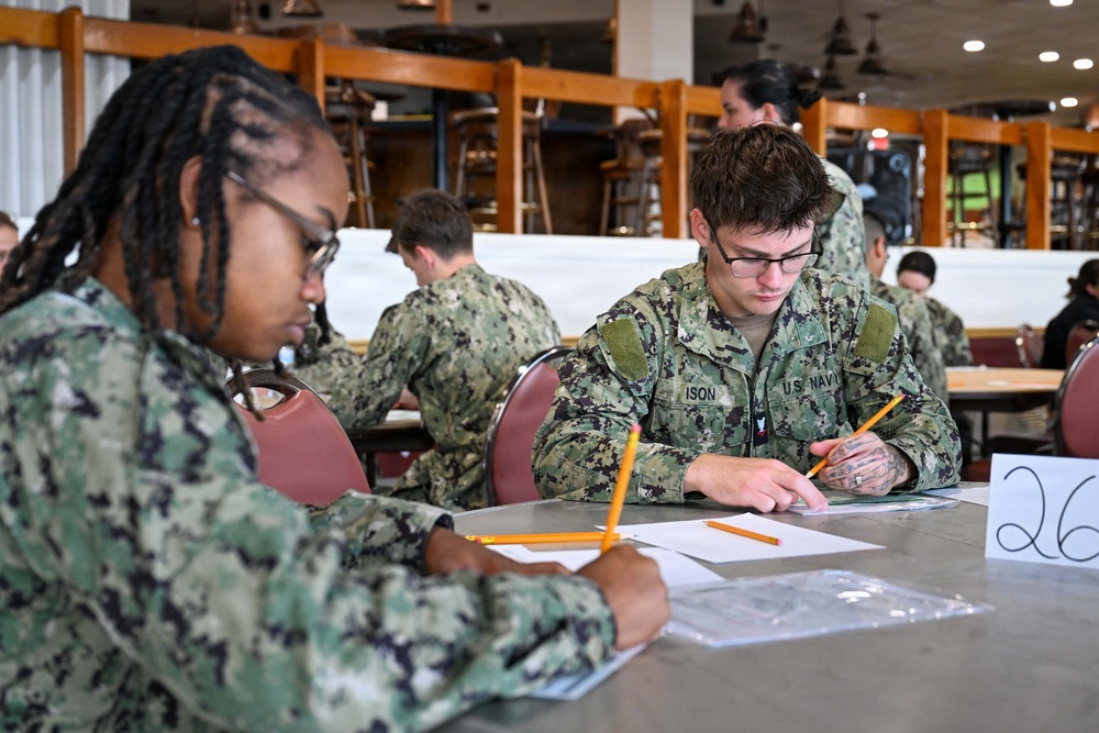 Multiple U.S. Navy personnel in NWU Type III camouflage uniforms are seated at tables in what appears to be a dining facility or classroom setting, completing written work. The foreground shows two sailors focused on paperwork, with one identified by name tape as 'ISON.' Additional sailors are visible in the background engaged in the same activity.
