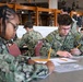 Multiple U.S. Navy personnel in NWU Type III camouflage uniforms are seated at tables in what appears to be a dining facility or classroom setting, completing written work. The foreground shows two sailors focused on paperwork, with one identified by name tape as 'ISON.' Additional sailors are visible in the background engaged in the same activity.