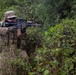 A soldier in full multicam camouflage gear is positioned in dense vegetation, aiming an assault rifle forward. The individual is equipped with tactical body armor, a plate carrier with magazine pouches, and optical sights mounted on the weapon. The setting appears to be a field training exercise or operational patrol in a wooded or brushy environment.