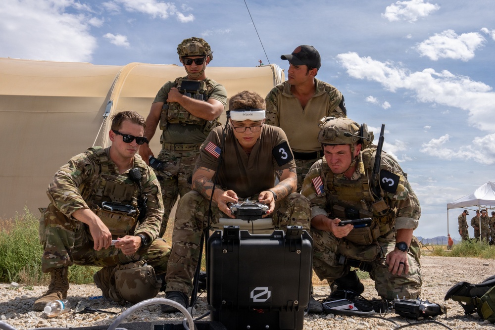 A group of five military personnel in camouflage uniforms are gathered around a Zeiss-branded equipment case in an outdoor desert training environment. The central figure is wearing FPV (First Person View) goggles and operating a drone controller, suggesting a drone operation or training exercise. Several team members wear the number '3' patch, indicating team designation, and U.S. flag patches are visible on uniforms.