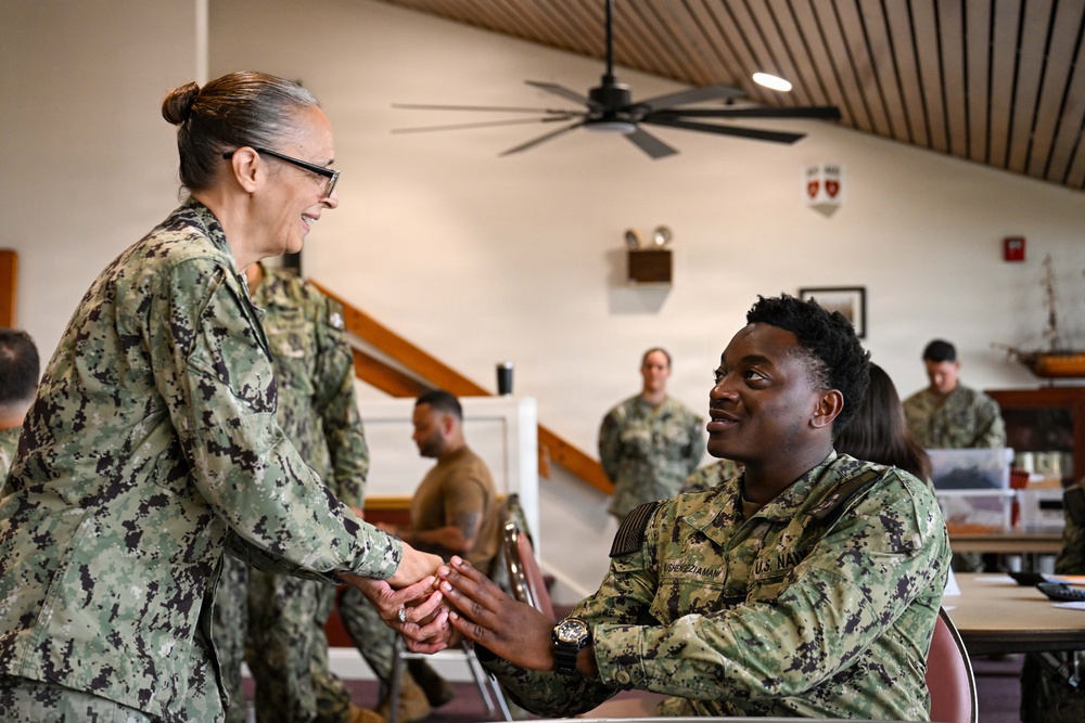 Two U.S. Navy personnel in NWU Type III (digital camouflage) uniforms are engaged in a handshake inside what appears to be a military facility or meeting hall. A senior female service member is shaking hands with a seated male sailor, suggesting a formal recognition or greeting interaction. Additional military personnel are visible in the background, some seated at tables and others standing.