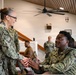Two U.S. Navy personnel in NWU Type III (digital camouflage) uniforms are engaged in a handshake inside what appears to be a military facility or meeting hall. A senior female service member is shaking hands with a seated male sailor, suggesting a formal recognition or greeting interaction. Additional military personnel are visible in the background, some seated at tables and others standing.