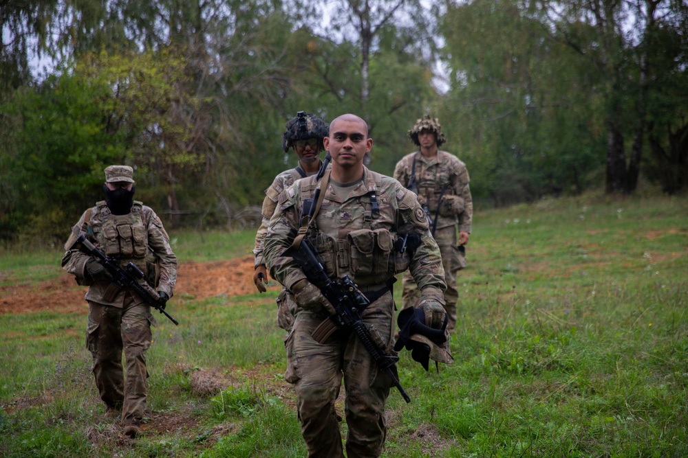 Four military personnel in camouflage uniforms are walking through an open grassy field toward the camera, with a wooded tree line in the background. The lead soldier is carrying an assault rifle at his side and holding a helmet in his other hand. The setting appears to be a training exercise or patrol in a field environment.