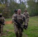 Four military personnel in camouflage uniforms are walking through an open grassy field toward the camera, with a wooded tree line in the background. The lead soldier is carrying an assault rifle at his side and holding a helmet in his other hand. The setting appears to be a training exercise or patrol in a field environment.