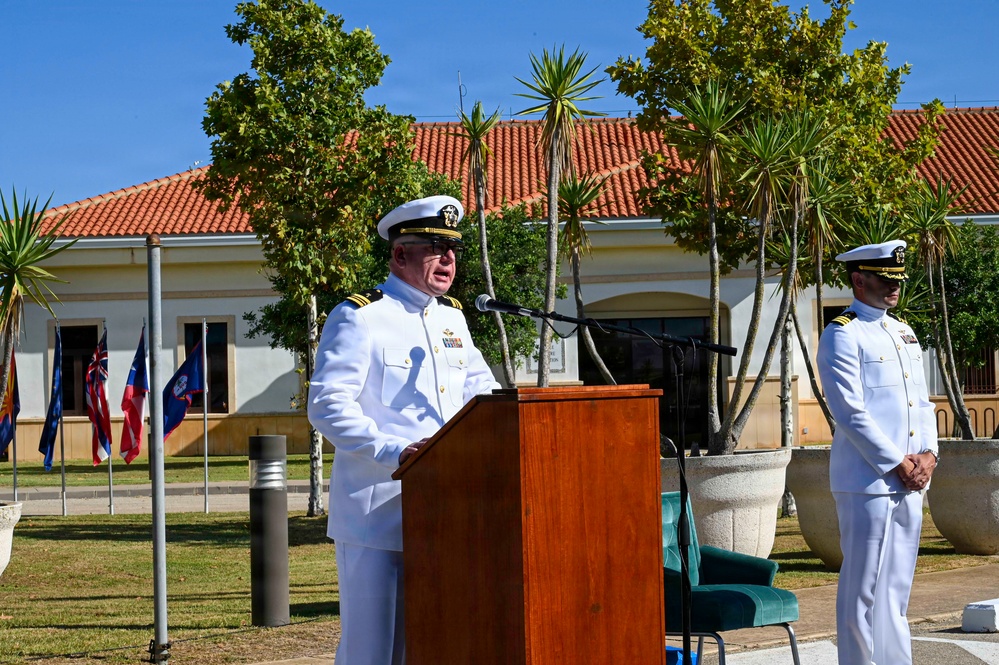 Two U.S. Navy officers in white dress uniforms are participating in a formal outdoor ceremony. One officer is speaking at a wooden podium equipped with a microphone, while a second officer stands at attention to the right. The setting appears to be a military installation with a Mediterranean-style building, multiple flags, and manicured grounds visible in the background.