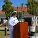 Two U.S. Navy officers in white dress uniforms are participating in a formal outdoor ceremony. One officer is speaking at a wooden podium equipped with a microphone, while a second officer stands at attention to the right. The setting appears to be a military installation with a Mediterranean-style building, multiple flags, and manicured grounds visible in the background.