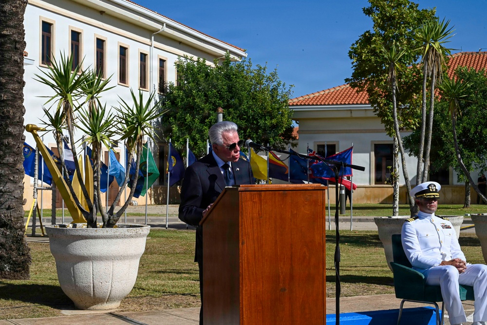 An outdoor formal ceremony is taking place at what appears to be a military or government installation. A civilian man in a dark suit is delivering a speech at a wooden podium with microphones, while a naval officer in white dress uniform is seated nearby. Multiple international flags are displayed in the background, suggesting a multinational or diplomatic event.