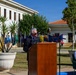 An outdoor formal ceremony is taking place at what appears to be a military or government installation. A civilian man in a dark suit is delivering a speech at a wooden podium with microphones, while a naval officer in white dress uniform is seated nearby. Multiple international flags are displayed in the background, suggesting a multinational or diplomatic event.