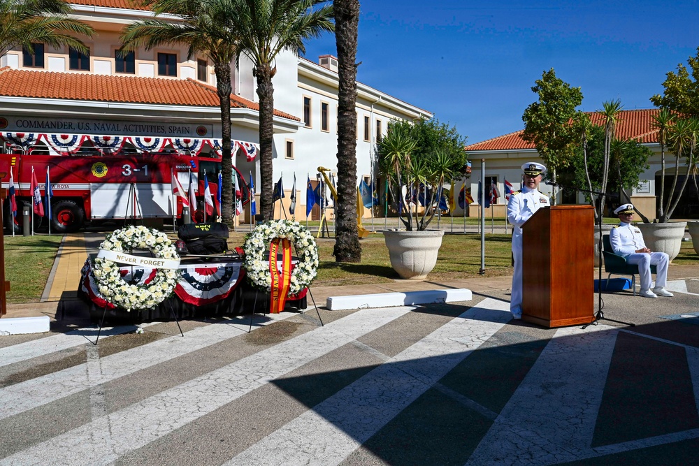 A memorial ceremony is taking place at Commander, U.S. Naval Activities, Spain, likely a 9/11 remembrance event based on the 'Never Forget' wreath and patriotic decorations. A naval officer in white dress uniform is delivering remarks at a wooden podium, while another officer is seated nearby. Two floral wreaths are displayed on stands in front of a fire truck numbered '3-1' adorned with red, white, and blue bunting.