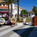 A memorial ceremony is taking place at Commander, U.S. Naval Activities, Spain, likely a 9/11 remembrance event based on the 'Never Forget' wreath and patriotic decorations. A naval officer in white dress uniform is delivering remarks at a wooden podium, while another officer is seated nearby. Two floral wreaths are displayed on stands in front of a fire truck numbered '3-1' adorned with red, white, and blue bunting.