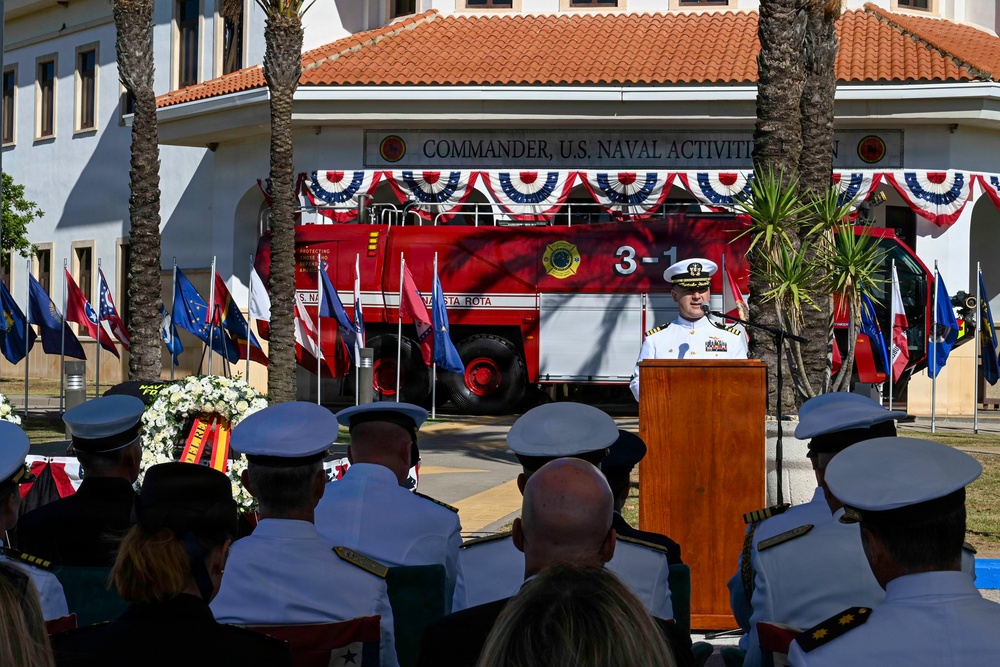A formal military ceremony is taking place in front of a building marked 'Commander, U.S. Naval Activities,' likely at Naval Station Rota, Spain. A naval officer in white dress uniform is speaking at a wooden podium before an audience of military personnel in white uniforms. A red fire truck labeled '3-1' and 'Naval Station Rota' is prominently displayed behind the speaker, decorated with red, white, and blue bunting.