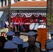 A formal military ceremony is taking place in front of a building marked 'Commander, U.S. Naval Activities,' likely at Naval Station Rota, Spain. A naval officer in white dress uniform is speaking at a wooden podium before an audience of military personnel in white uniforms. A red fire truck labeled '3-1' and 'Naval Station Rota' is prominently displayed behind the speaker, decorated with red, white, and blue bunting.