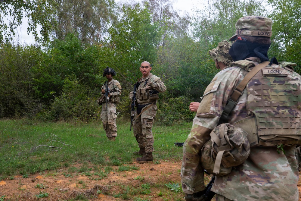 Three military personnel in MultiCam/OCP camouflage uniforms are gathered in a clearing near a treeline. The soldier in the foreground, identified by name tape as 'LOONEY,' is facing two other soldiers who appear to be receiving instruction or a briefing. The setting appears to be a field training exercise in a wooded outdoor environment.