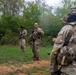 Three military personnel in MultiCam/OCP camouflage uniforms are gathered in a clearing near a treeline. The soldier in the foreground, identified by name tape as 'LOONEY,' is facing two other soldiers who appear to be receiving instruction or a briefing. The setting appears to be a field training exercise in a wooded outdoor environment.