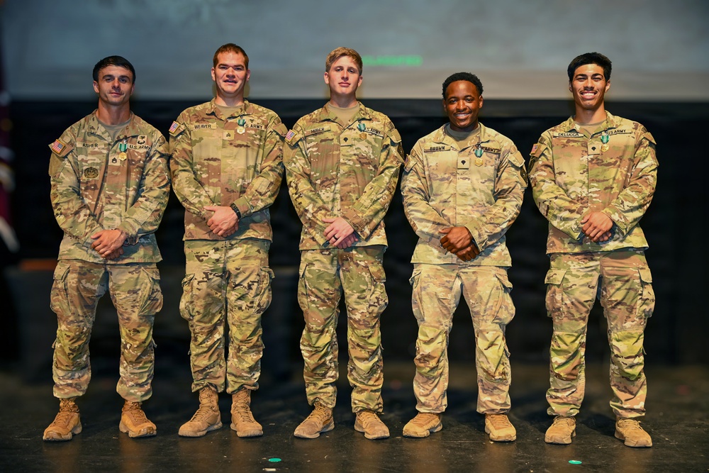 Five U.S. Army soldiers in OCP (Operational Camouflage Pattern) uniforms are posed together on a stage, appearing to be at an awards or recognition ceremony. Each soldier is wearing medals or awards on their chest, suggesting this is a formal military recognition event. Name tapes visible on their uniforms include Asher, Weaver, Moler, Brown, and Delucca.