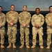 Five U.S. Army soldiers in OCP (Operational Camouflage Pattern) uniforms are posed together on a stage, appearing to be at an awards or recognition ceremony. Each soldier is wearing medals or awards on their chest, suggesting this is a formal military recognition event. Name tapes visible on their uniforms include Asher, Weaver, Moler, Brown, and Delucca.