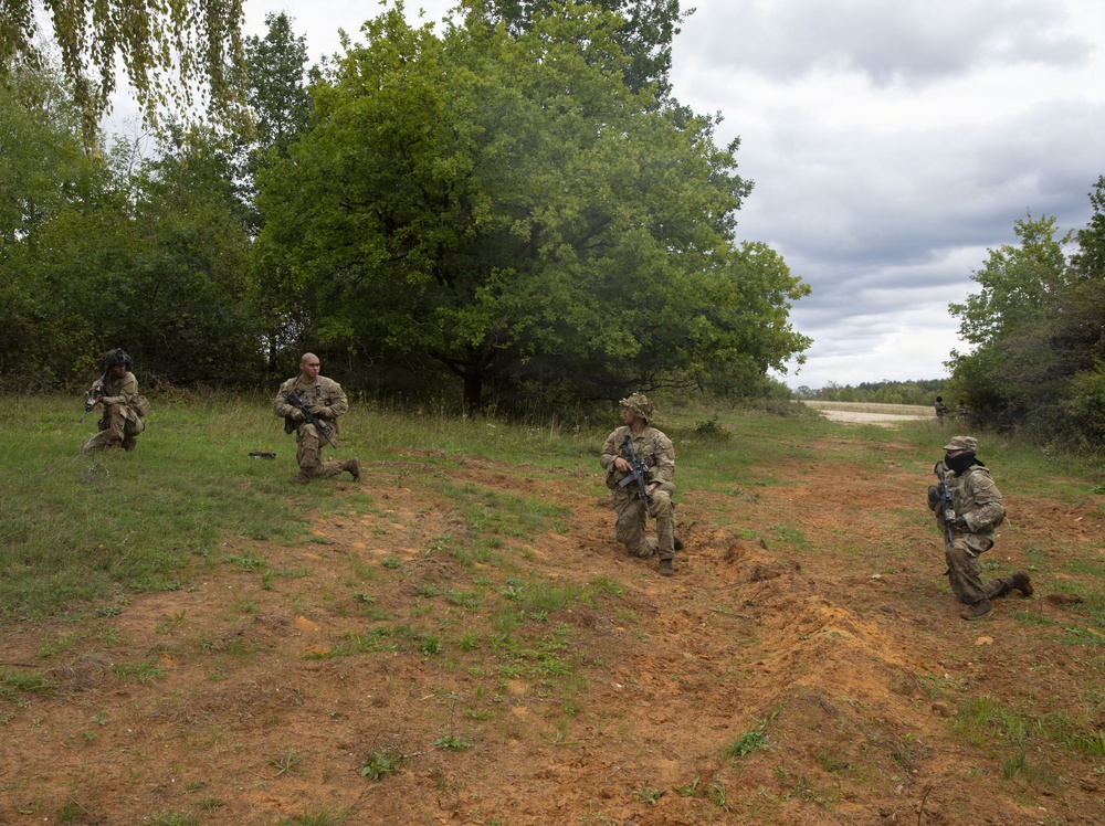 Four military personnel in camouflage uniforms are positioned in a kneeling tactical formation across an open grassy field with disturbed earth. They are armed with rifles and appear to be conducting a field training exercise or tactical movement drill. A dirt road and treeline are visible in the background under an overcast sky.