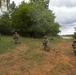 Four military personnel in camouflage uniforms are positioned in a kneeling tactical formation across an open grassy field with disturbed earth. They are armed with rifles and appear to be conducting a field training exercise or tactical movement drill. A dirt road and treeline are visible in the background under an overcast sky.