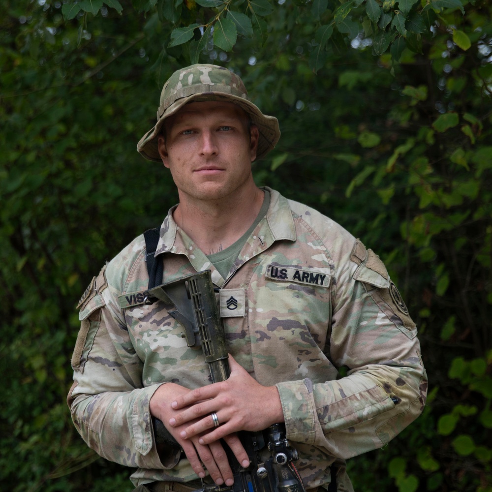 A U.S. Army soldier in full MultiCam (OCP) camouflage uniform poses for a portrait against a green foliage background. He is wearing a boonie hat and holding a rifle, with rank insignia and name tape visible on his uniform. The image appears to be a professional or semi-professional military portrait.