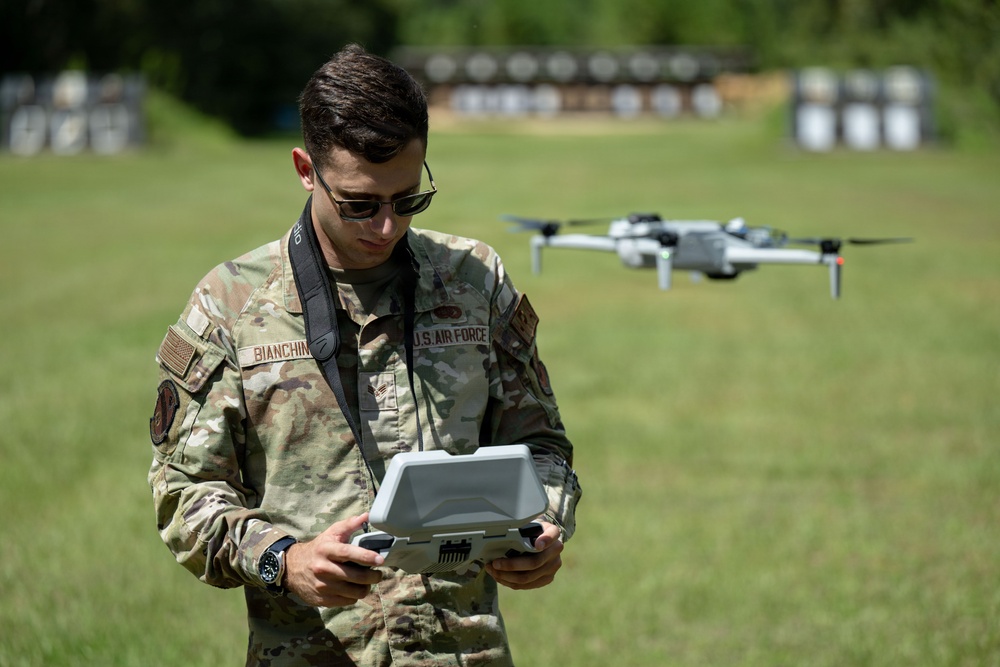 A U.S. Air Force service member identified by name tape as 'BIANCHINI' operates a small quadcopter drone using a handheld remote controller with an integrated screen. The airman is wearing OCP (Operational Camouflage Pattern) uniform and sunglasses, standing on a grass field with what appears to be a shooting range target backdrop in the background. A DJI-style consumer or commercial drone is visibly airborne behind him.