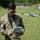 A U.S. Air Force service member identified by name tape as 'BIANCHINI' operates a small quadcopter drone using a handheld remote controller with an integrated screen. The airman is wearing OCP (Operational Camouflage Pattern) uniform and sunglasses, standing on a grass field with what appears to be a shooting range target backdrop in the background. A DJI-style consumer or commercial drone is visibly airborne behind him.