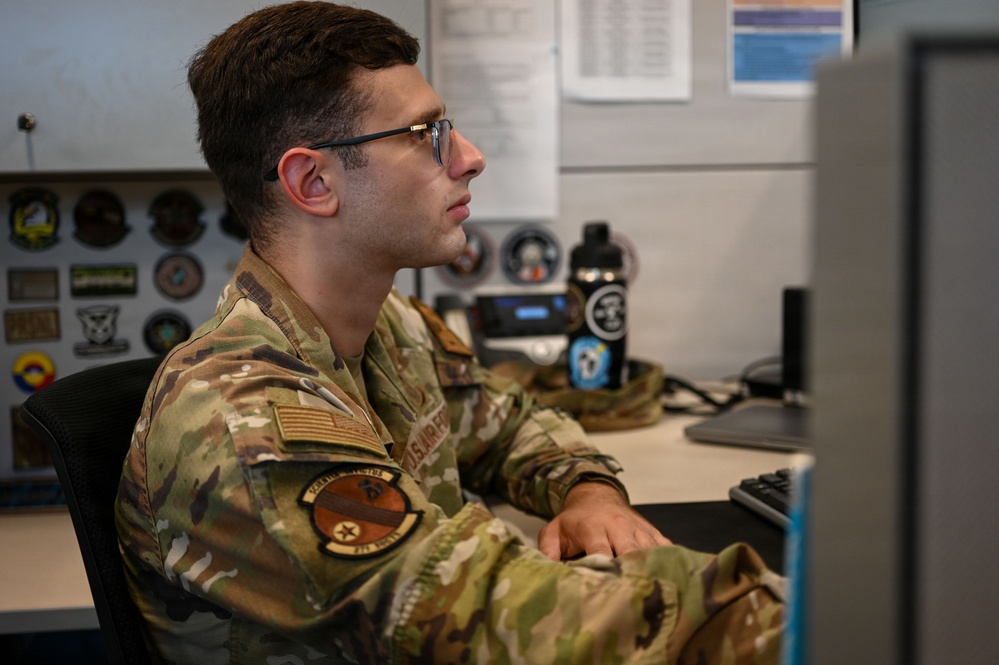 A young male U.S. Air Force service member in OCP (Operational Camouflage Pattern) uniform is seated at a desk working at a computer workstation. He is wearing glasses and has a U.S. flag patch and a squadron patch visible on his left sleeve. The setting appears to be a military office or operations center with unit patches and insignia displayed on the wall behind him.