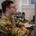 A young male U.S. Air Force service member in OCP (Operational Camouflage Pattern) uniform is seated at a desk working at a computer workstation. He is wearing glasses and has a U.S. flag patch and a squadron patch visible on his left sleeve. The setting appears to be a military office or operations center with unit patches and insignia displayed on the wall behind him.