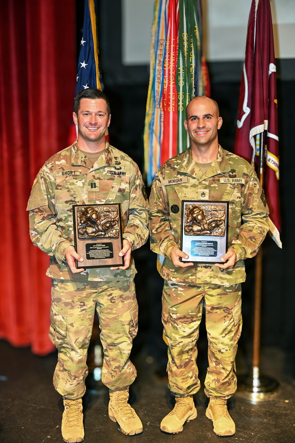 Two U.S. Army soldiers in OCP (Operational Camouflage Pattern) uniforms are posing on a stage holding identical bronze relief plaques/awards. The soldier on the left has the name tape 'SHORT' and appears to hold the rank of Captain, while the soldier on the right has the name tape 'MORGAN' and wears Staff Sergeant rank insignia. They are standing in front of multiple military unit flags and a U.S. flag in what appears to be a formal awards ceremony.