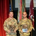 Two U.S. Army soldiers in OCP (Operational Camouflage Pattern) uniforms are posing on a stage holding identical bronze relief plaques/awards. The soldier on the left has the name tape 'SHORT' and appears to hold the rank of Captain, while the soldier on the right has the name tape 'MORGAN' and wears Staff Sergeant rank insignia. They are standing in front of multiple military unit flags and a U.S. flag in what appears to be a formal awards ceremony.