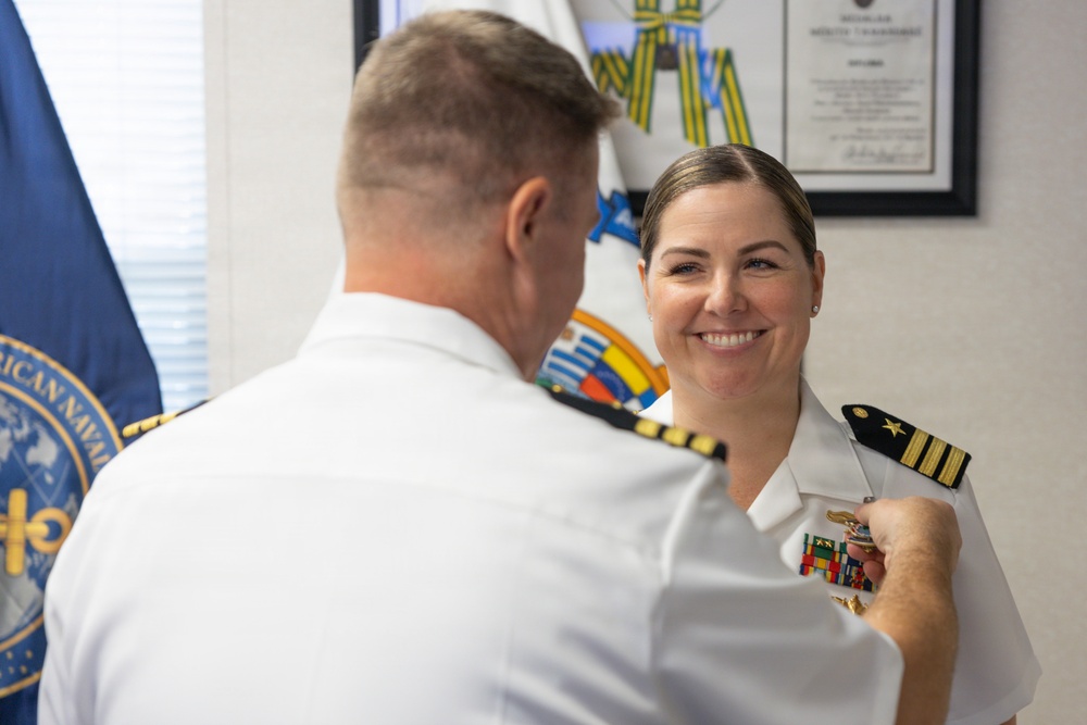 A military pinning ceremony is taking place between two naval officers in white dress uniforms. A male officer, seen from behind, is pinning a medal or insignia onto the uniform of a smiling female officer. The setting appears to be an official military office with flags and a framed certificate visible in the background.