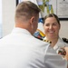 A military pinning ceremony is taking place between two naval officers in white dress uniforms. A male officer, seen from behind, is pinning a medal or insignia onto the uniform of a smiling female officer. The setting appears to be an official military office with flags and a framed certificate visible in the background.