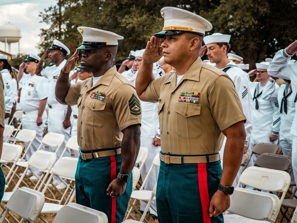 A military ceremony featuring United States Marine Corps personnel in the foreground saluting, wearing Service Charlie uniforms with green trousers and red blood stripes. Behind them, a large formation of U.S. Navy sailors in white dress uniforms are also saluting. The setting appears to be an outdoor ceremonial event, likely a change of command, retirement, or memorial service.