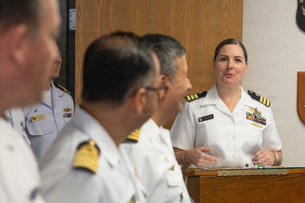 A female naval officer in white dress uniform is speaking at a podium during what appears to be an official military ceremony or briefing. She is addressing a group of male naval officers seated or standing in the foreground, who are wearing white dress uniforms with gold insignia. The setting appears to be an indoor military facility or conference room.