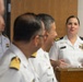 A female naval officer in white dress uniform is speaking at a podium during what appears to be an official military ceremony or briefing. She is addressing a group of male naval officers seated or standing in the foreground, who are wearing white dress uniforms with gold insignia. The setting appears to be an indoor military facility or conference room.